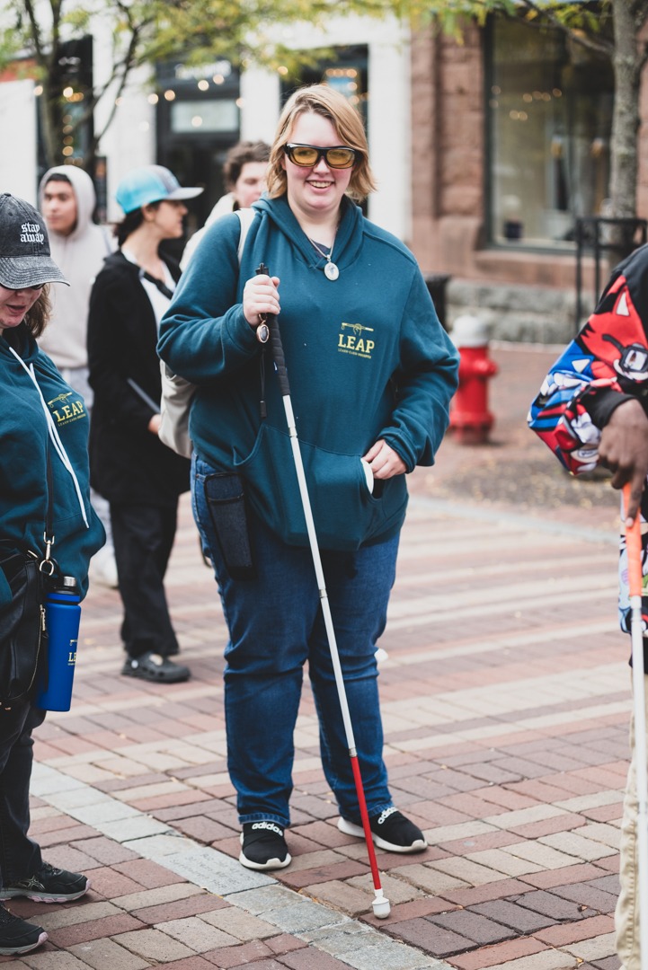 A smiling individual with visual impairment is featured prominently in the foreground, navigating a brick-paved sidewalk with a white cane. They are wearing yellow-tinted glasses, a dark blue hoodie with the 'LEAP' logo in gold, denim jeans, and black sneakers with white soles. A water bottle in a holder and a small pouch are attached to their belt loop. In the softly focused background, there are several people who appear to be part of a group or public event, suggesting a communal outdoor setting.