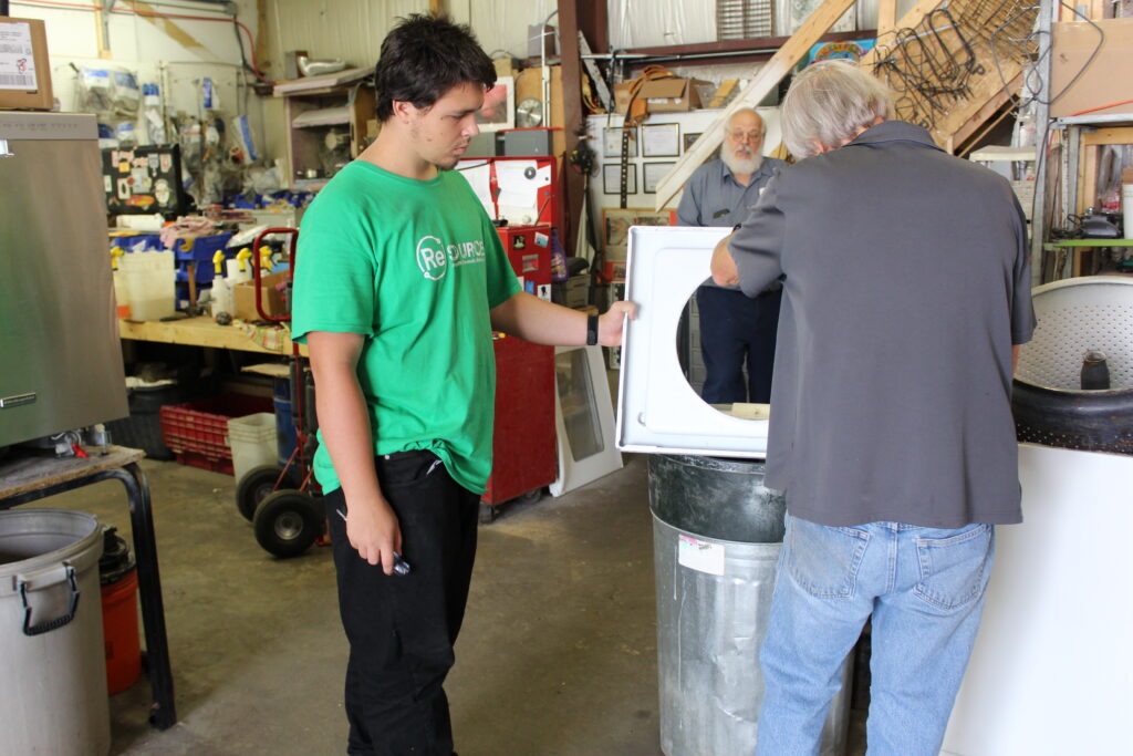 ReSOURCE Appliance Repair workers troubleshoot a large appliance. One holds the dryer door open while the other inspects the machine, seeking assistance. Various tools and equipment are visible in the background, ready for use.