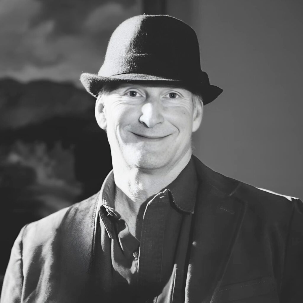 Eric Weeber wearing a dark fedora and jacket stands smiling, embodying the spirit of sustainable living. The black and white photo, with its blurred background, highlights his cheerful expression and classic attire.