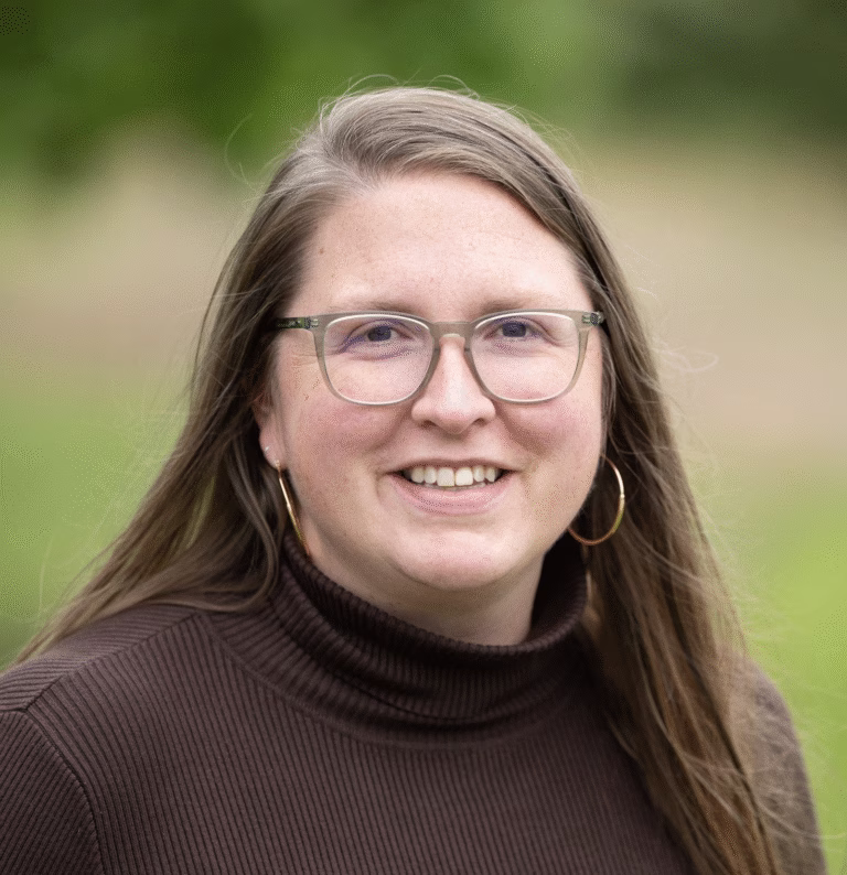 A woman with long brown hair, wearing glasses, hoop earrings, and a dark brown turtleneck sweater, smiles outdoors with a blurred green ReSOURCE background.