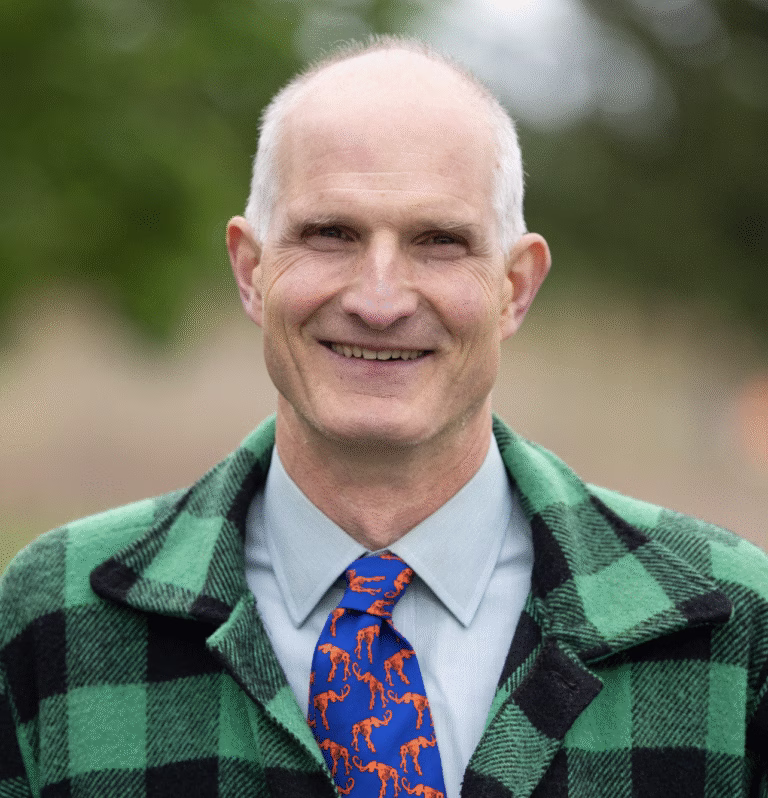 Tom Longstreth, wearing a green and black plaid jacket, a light blue shirt, and a blue tie with orange mastedons, stands outdoors amid greenery, representing ReSOURCE with confidence and warmth.