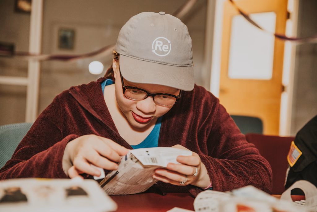 A ReSOURCE LEAP trainee wearing glasses and a gray cap with a “Re” logo examines a small box closely while sitting at a table, appearing focused—perhaps testing something for LEAP students—in an indoor space with blurred background details.