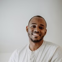 Ibby Wilson with short hair and a trimmed beard is smiling at the camera. He is wearing a light-colored, button-up shirt and stands in front of a plain background, embodying the approachable spirit of the ReSOURCE team.