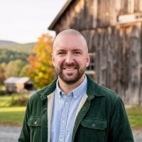 Tom Johnston stands outside a rustic wooden barn, wearing a green jacket over a light blue shirt. Trees and hills are visible in the background.