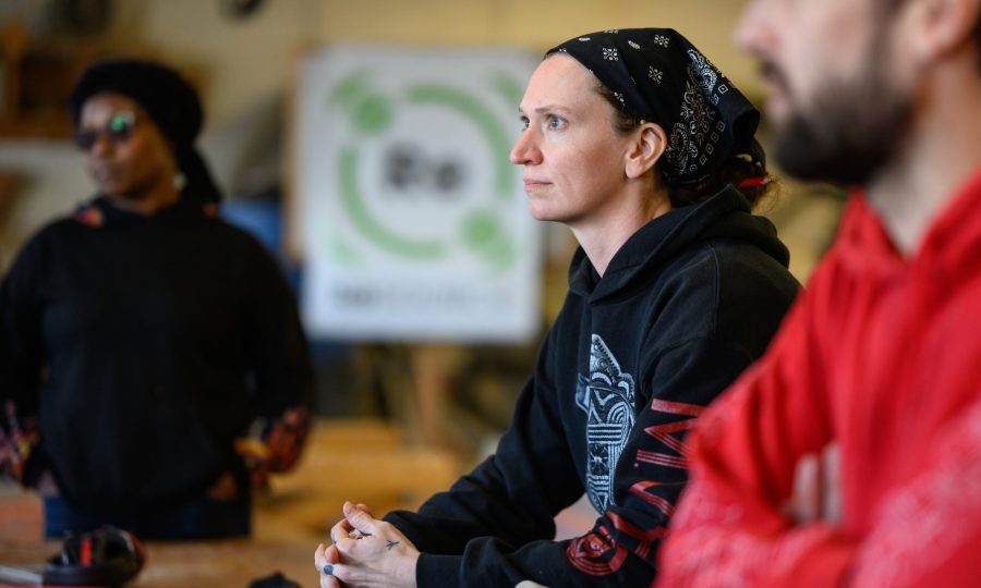 A ReSOURCE trainee in a black hoodie and bandana looks focused in a workshop, with two people nearby. A green and white ReSOURCE sign, promoting reuse, is blurred in the background.