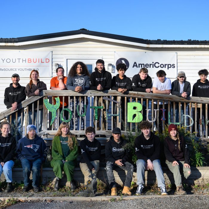 ReSOURCE YouthBuild Burlington Students and Staff pose on a ramp and walkway outside a building with "YouthBuild" and "AmeriCorps" banners, wearing casual clothes and hoodies, with greenery and handmade "YouthBuild" sign in front.