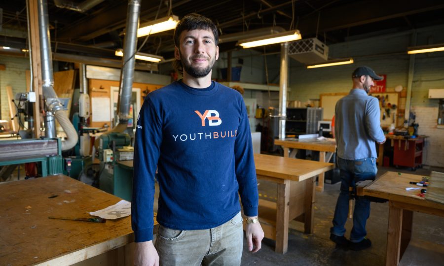 Noah Kass from ReSOURCE YouthBuild Burlington wearing a “YouthBuild” shirt stands and smiles in a woodworking shop at ReSOURCE, with wooden workbenches and tools around him. Another person works in the background, showcasing the hands-on spirit found at ReSOURCE.