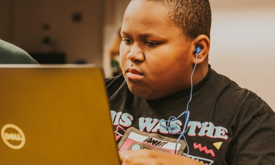 A young boy wearing a graphic T-shirt and blue earphones sits at a table, focused on working on a Dell laptop in a brightly lit indoor setting, showcasing the benefits of ReSOURCE and reuse in home improvement and learning spaces.