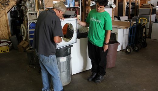 A ReSOURCE technician in a green shirt supervises while a colleague services a front-loading dryer, illustrating the large appliance sales and services available at ReSOURCE locations.