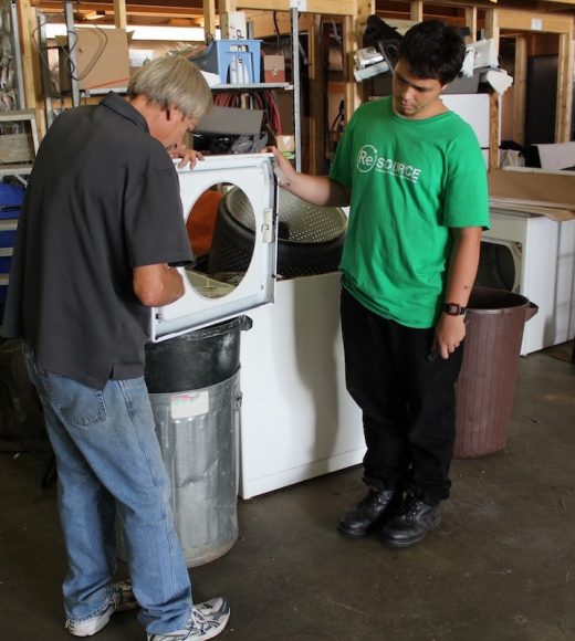 A ReSOURCE technician in a green shirt supervises while a colleague services a front-loading dryer, illustrating the large appliance sales and services available at ReSOURCE locations.