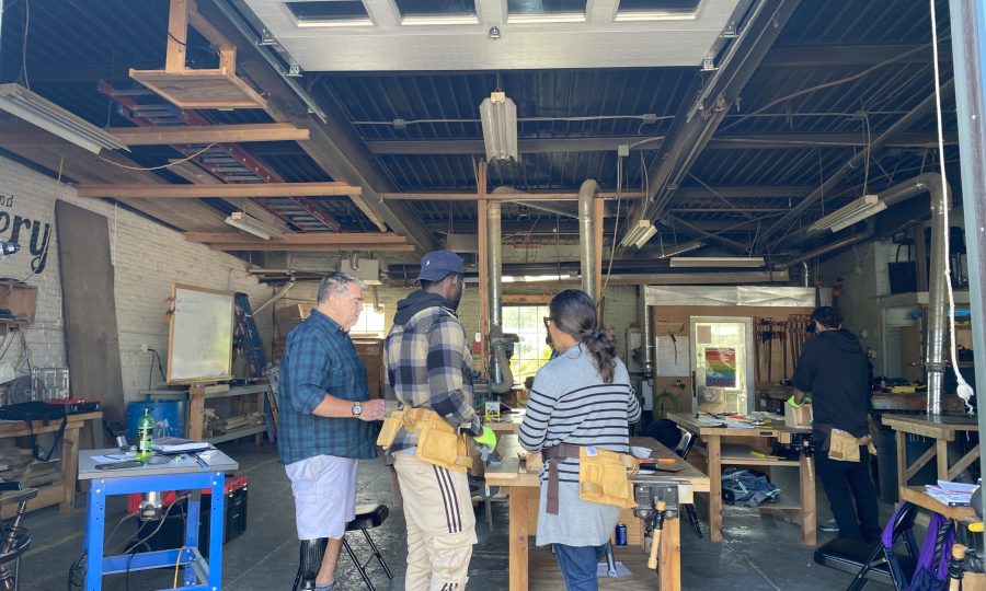 A group of trainees in a workshop stands around a large workbench. Equipped with tool belts, they engage in hands-on training, surrounded by woodworking tools and equipment.