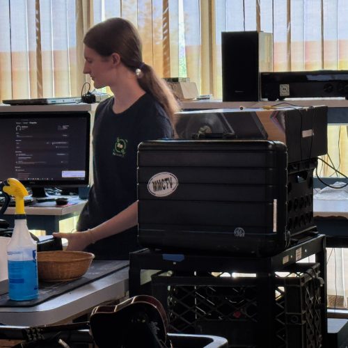 A ReSOURCE Employee stands near a desk with computer screens, representing Computer Services, alongside a spray bottle, a basket, and stacked black bins. Vertical blinds cover windows in the background, letting in sunlight.