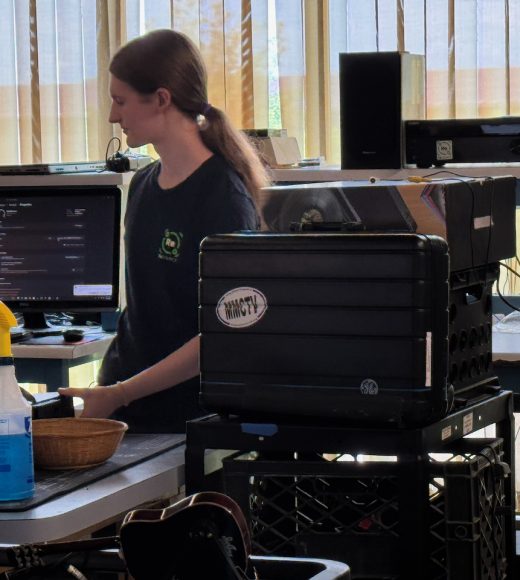 A ReSOURCE Employee stands near a desk with computer screens, representing Computer Services, alongside a spray bottle, a basket, and stacked black bins. Vertical blinds cover windows in the background, letting in sunlight.