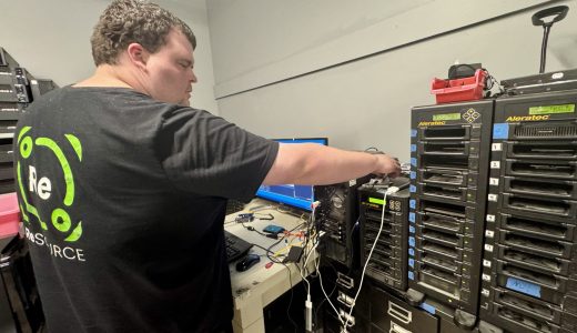 A ReSOURCE Employee stands in a tech room connecting cables to a computer tower, surrounded by electronics, monitors, and stacked hard drive duplicators—showcasing ReSOURCE’s commitment to sustainability through the mindful reuse and repurposing of technology.