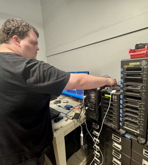 A ReSOURCE Employee stands in a tech room connecting cables to a computer tower, surrounded by electronics, monitors, and stacked hard drive duplicators—showcasing ReSOURCE’s commitment to sustainability through the mindful reuse and repurposing of technology.
