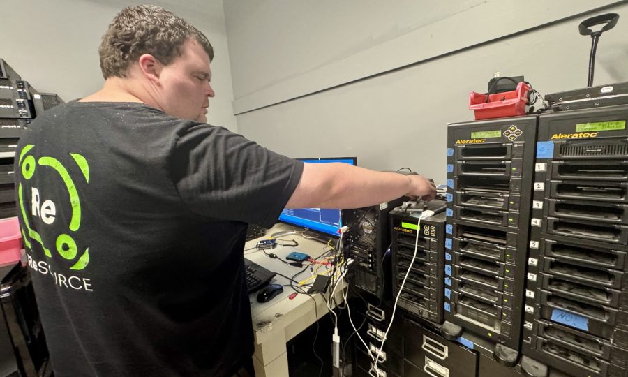A ReSOURCE Employee stands in a tech room connecting cables to a computer tower, surrounded by electronics, monitors, and stacked hard drive duplicators—showcasing ReSOURCE’s commitment to sustainability through the mindful reuse and repurposing of technology.