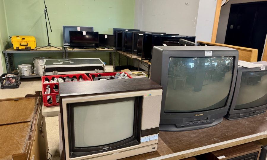 Several old televisions and monitors are displayed on tables in a store, alongside some audio equipment and a yellow stereo visible in the background. The setting appears to be a secondhand or thrift shop that may also offer computer sales and related computer services.