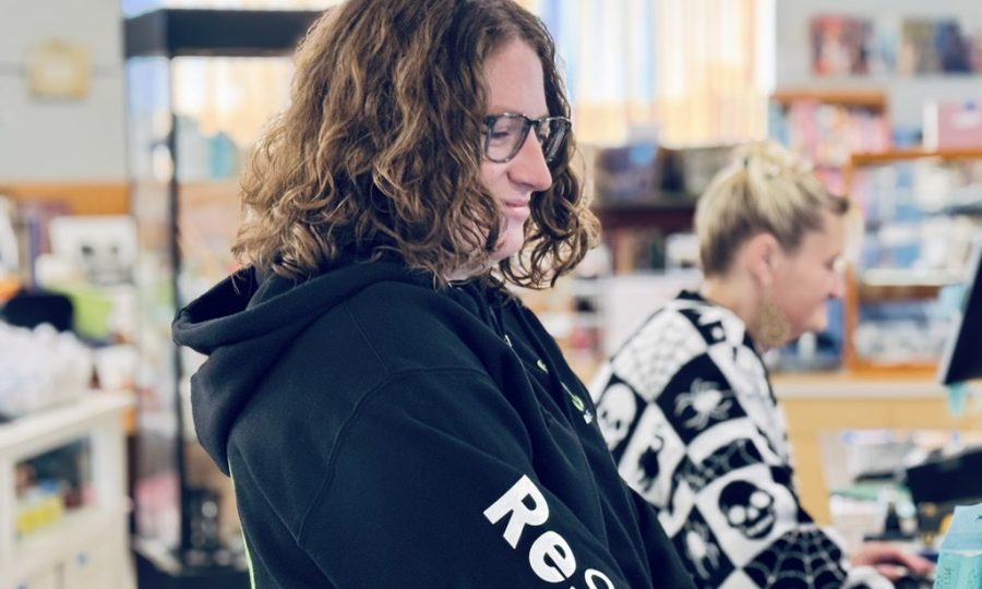 A ReSOURCE Employee smiles while working at a counter in what appears to be a ReSOURCE store or library. Another person in a black-and-white sweater is in the background, focused on a computer, creating an inviting Home Page atmosphere for visitors.