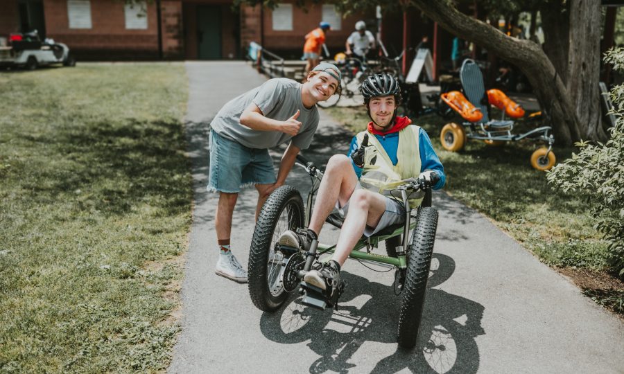 A young man wearing a helmet sits in an adaptive off-road trike on a paved path, while another person beside him smiles and gives a thumbs up. Trees and outdoor activity equipment are visible in the background.