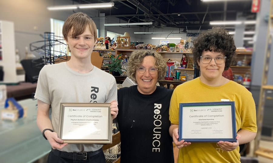 ReSOURCE Trainees stand indoors, smiling at the camera. The person in the middle wears a “ReSOURCE” shirt, highlighting their involvement in local training programs. The two people on either side hold framed certificates of completion, celebrating their achievements. Shelves and items are visible in the background.