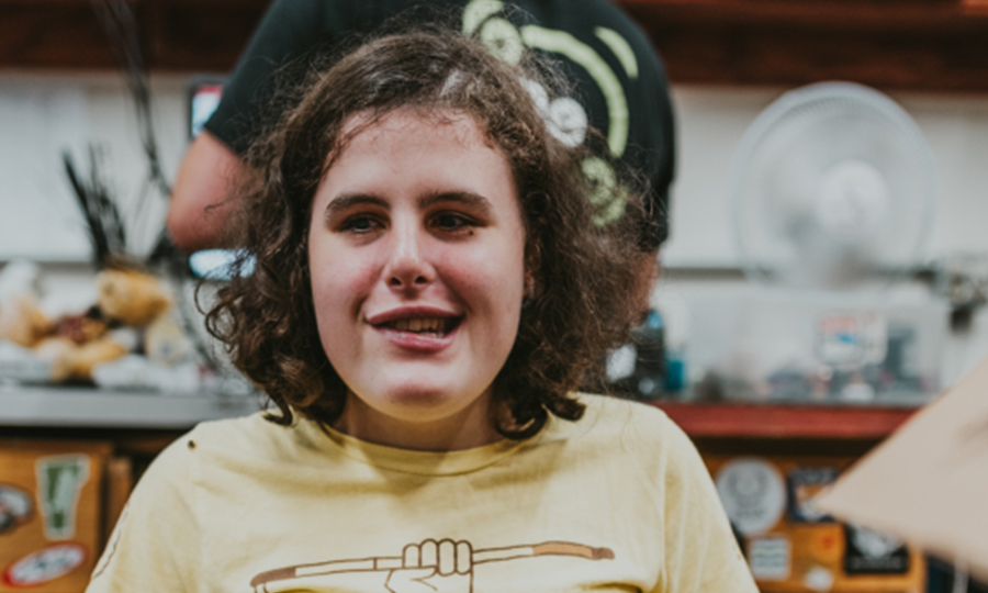 A young student with curly hair, wearing a yellow t-shirt with a graphic design, smiles indoors. The background shows shelves, a fan, and various objects, slightly out of focus—capturing the vibrant spirit of LEAP participants.