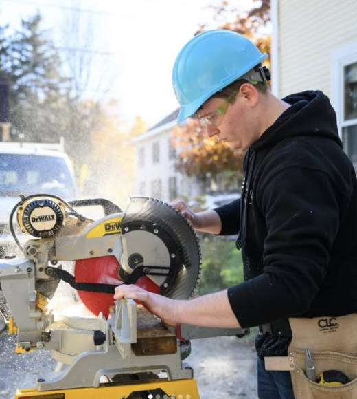 A ReSOURCE YouthBuild Student wearing a blue hard hat and safety glasses uses a Dewalt circular saw to cut wood outdoors, with sawdust flying and houses visible in the background.