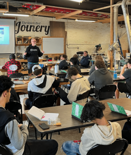 A group of YouthBuild students sits at tables in a workshop, attentively facing an instructor who is presenting information on a screen. The room contains various tools and equipment for woodworking or crafting.