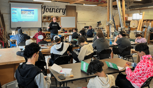 A group of YouthBuild students sits at tables in a workshop, attentively facing an instructor who is presenting information on a screen. The room contains various tools and equipment for woodworking or crafting.