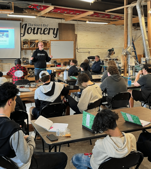 A group of YouthBuild students sits at tables in a workshop, attentively facing an instructor who is presenting information on a screen. The room contains various tools and equipment for woodworking or crafting.