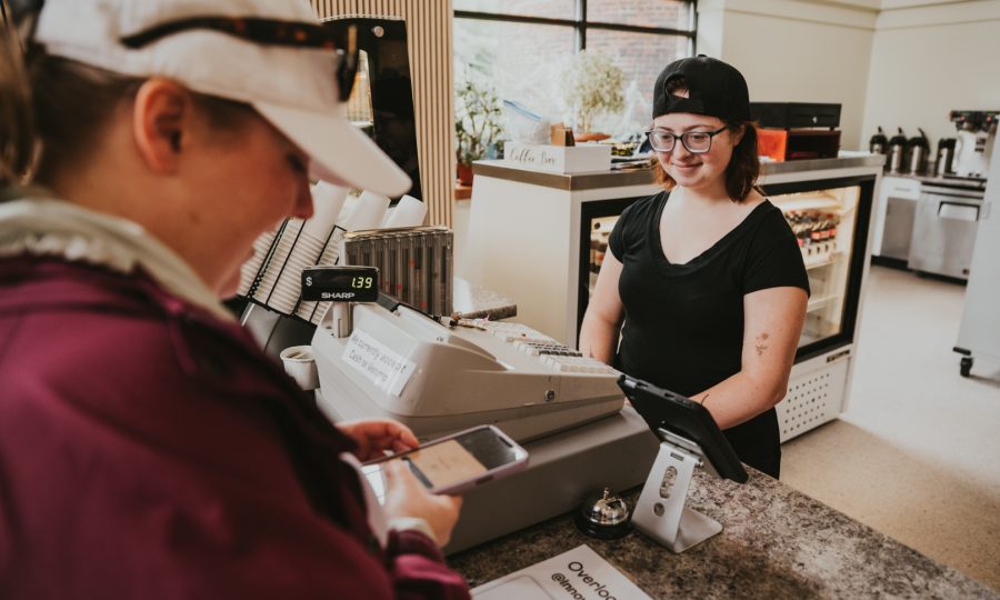 At LEAP Innovation Cafe, a customer is sporting a white cap and maroon jacket uses a smartphone at the cafe's counter, seemingly browsing the ReSOURCE Home Page. The cashier, wearing glasses and a black cap, smiles warmly from behind the register, while a display case and coffee machines add to the cozy ambiance.